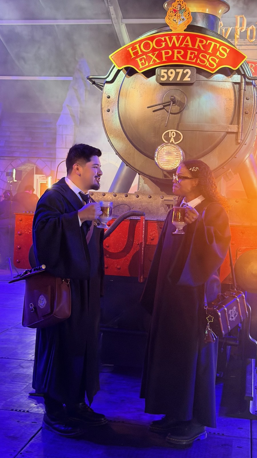 Two guests in Hogwarts robes holding Butterbeer glasses in front of the Hogwarts Express 5972 engine