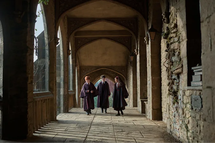 Harry, Ron and Hermione in Hogwarts robes walking a castle corridor in the HBO Harry Potter series